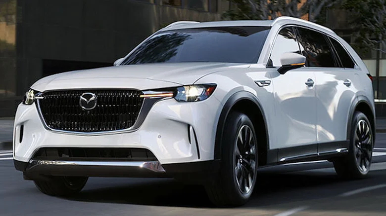 A white CX-90 driving past trees and a glossy building in a city