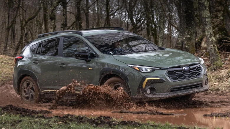 An olive green Subaru Crosstrek splashing through mud in a wooded area.