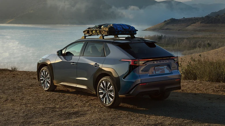 A shiny blue Subaru Solterra with gear in a roof rack, parked next to a lake.